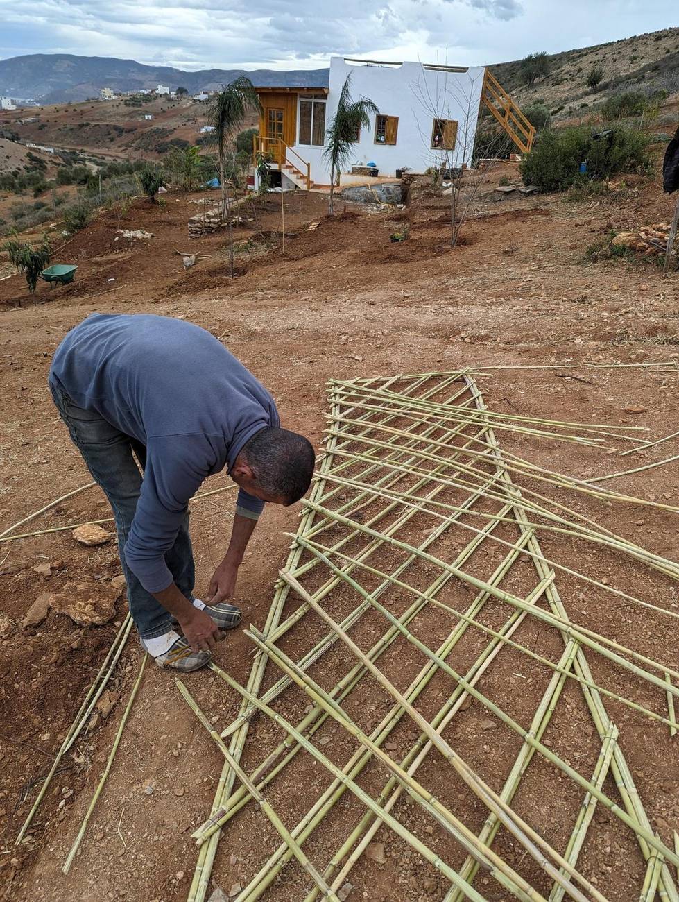 A craftsman weaving a cane and reed lattice by hand, with a bungalow in the background